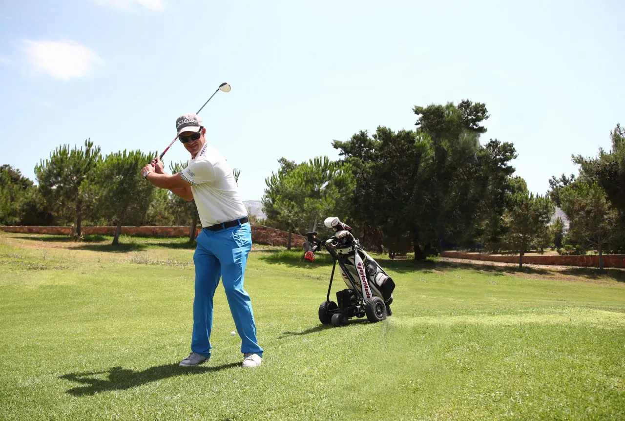 A golfer swinging on the fairway beside a golf trolley